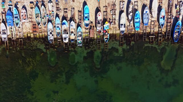 Many small boats are docked in a row at a wooden pier beside clear green water. The scene is under bright sunlight in a coastal region.