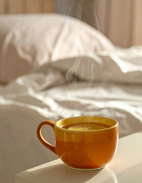Steaming orange ceramic cup of lemon tea on a bedside table with an unmade bed behind, for sick day and cold season