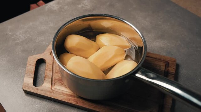 Peeled raw potatoes soaking in water inside a stainless steel saucepan on a wooden cutting board on a kitchen countertop