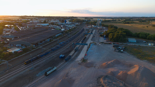 Aerial view of Wellingborough sidings freight terminal at sunset with freight trains on tracks, industrial warehouses, and piles of aggregate in Wellingborough, England, United Kingdom.