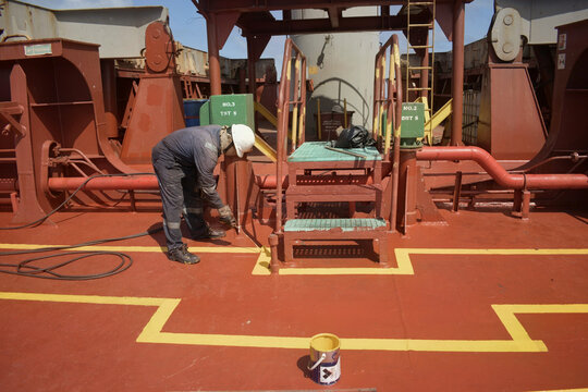 painting of yellow safety walkway boundary lines on the reddish deck of a cargo ship during vessel maintenance at sea