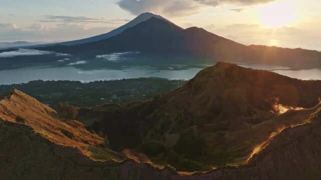 Aerial view drone flight over active Mount Batur caldera with steam vents, lake Batur, and the majestic Mount Agung volcano creating a serene Balinese landscape during colorful morning sunrise
