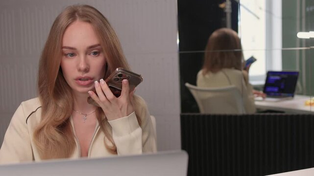 Female candidate communicates via phone with colleague visible in reflection. Caucasian woman engaged in professional remote interview with colleague reflected in glass