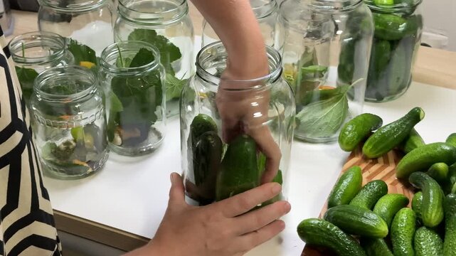 Multiple hands handle glass jars containing various types of green vegetables such as cucumbers and zucchini, suggesting a food preparation or storage activity set up on a white surface