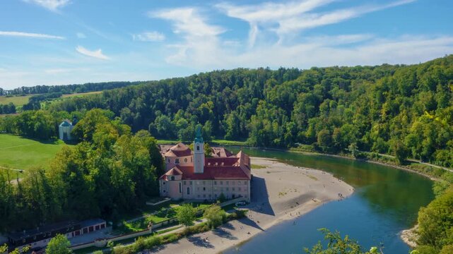 Timelapse aerial video of Weltenburg Abbey on the Danube River in Bavaria, Germany, surrounded by lush forests, riverside landscape and a scenic sandy shore