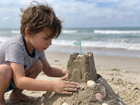 A little boy in a striped shirt carefully adds seashells to a sandcastle by the surf. Concept of seaside creativity and childhood play.