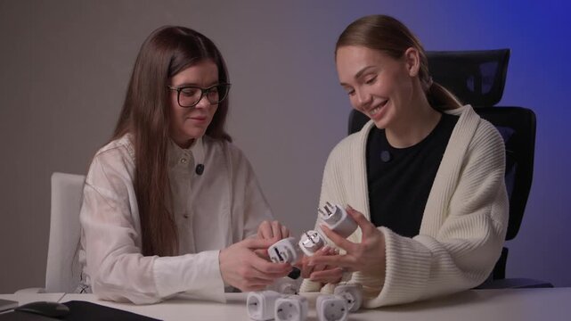 Caucasian women assess smart socket functionality carefully. Two ladies of Caucasian descent collaboratively inspect and compare smart socket models on studio desk