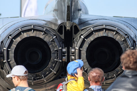 A detailed rear-view photo of a twin-engine military jet's exhaust nozzles with spectators, including a child in a blue cap, admiring the aircraft at an air show.