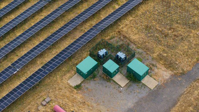 Aerial view of a solar farm featuring rows of photovoltaic panels alongside green battery storage units and electrical transformers in Bedford, England, United Kingdom.
