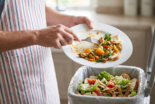 Person scraping leftover food from plate into kitchen trash bin after meal showing household food waste.