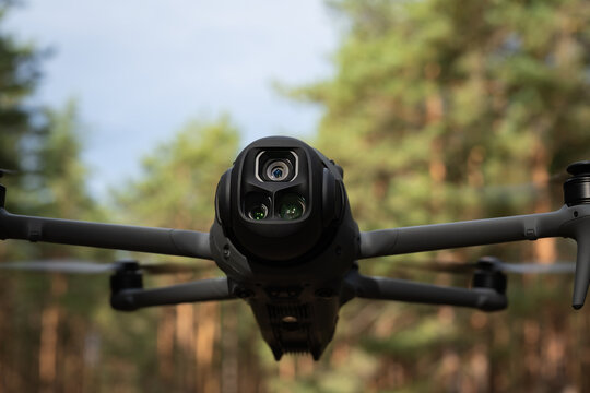 Close-up front view of a quadcopter drone with triple camera system hovering outdoors in a sunny pine forest during daylight.