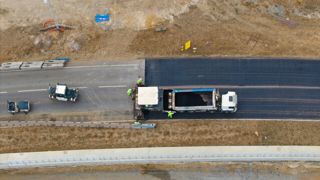Aerial view of road construction at Black Cat Roundabout with asphalt paving machine, dump truck, and workers in high-visibility gear in Chawston, England, United Kingdom.