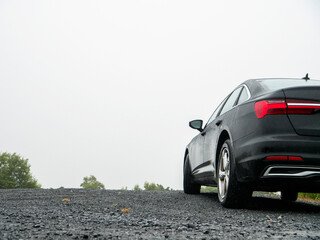 Black luxury sedan parked on gravel road. Misty sky above. Trees visible in distance. Car appears still and quiet. Suitable for travel, automotive, or landscape themes. Trip with comfort. © mark_gusev