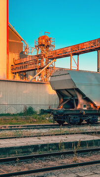 Industrial freight hopper car on tracks next to processing plant machinery illuminated by warm sunlight. Concept of heavy industry, freight logistics and material transport.