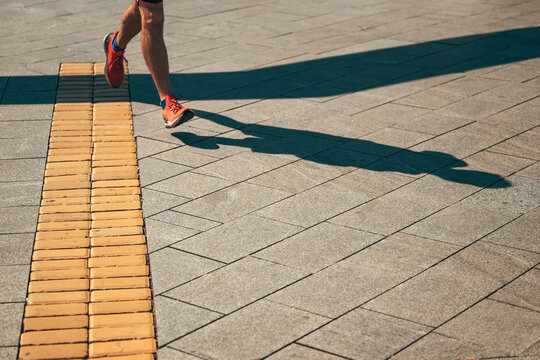 Dynamic high angle view of a runner's legs and shadow on urban pavement in strong sunlight. Concept of outdoor fitness, morning workouts, marathon running, and active lifestyle.