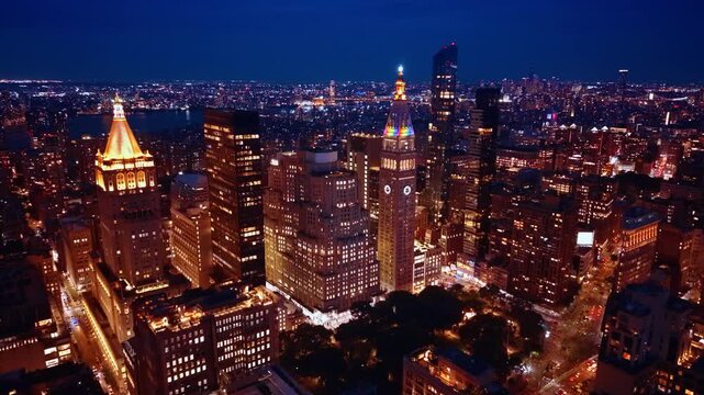 Madison Square Park and surrounding towers at night. Lit streets border a dark tree canopy beneath office towers and a glowing clock face.