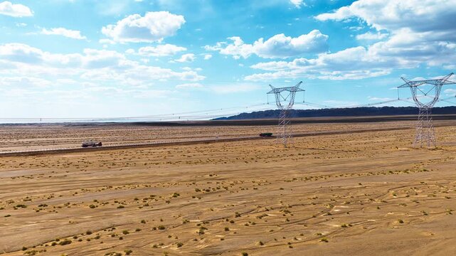 High-voltage transmission towers stand prominently in a vast desert landscape under a clear blue sky filled with white clouds.
