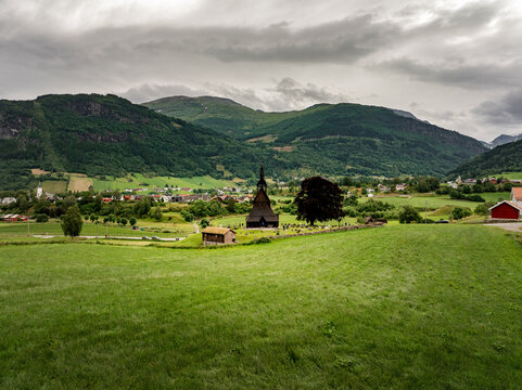 Aerial view of Hopperstad Stave Church set in green fields with a historic stone wall and tree, backed by the village of Vik i Sogn and mountains in Vik i Sogn, Vestland, Norway.