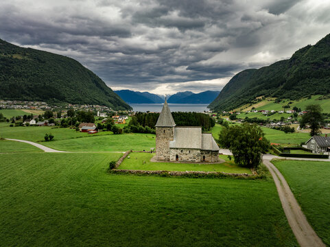 Aerial view of Hove Church surrounded by lush green fields and stone walls, with mountains and a fjord in the background in Vik i Sogn, Vestland, Norway.