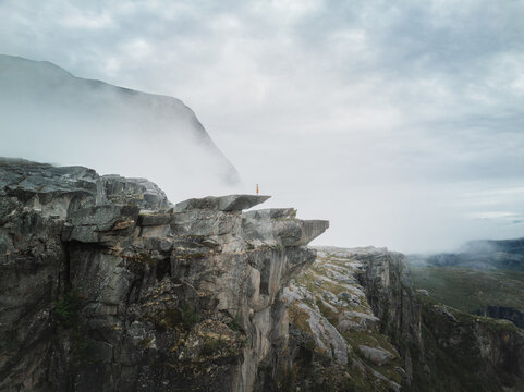 Aerial view of a person standing on the edge of the rocky Hellemojuvet canyon surrounded by mist and dramatic mountains in Hellmobotn, Nordland, Norway.