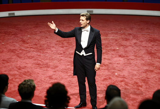 Entertainment and performance, circus ringmaster in tuxedo presenting show to audience in red arena with spotlight and theatrical atmosphere for event