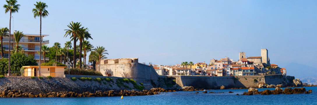 Panoramic view of Antibes Old Town with ancient ramparts, Grimaldi Castle tower, colorful houses, palm trees and rocky Mediterranean coast, French Riviera, France