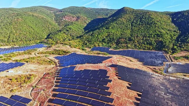 Rows of solar panels stretch across the land in a mountainous region. The project aims to produce clean energy. The sky is clear and the sun is shining on the panels.