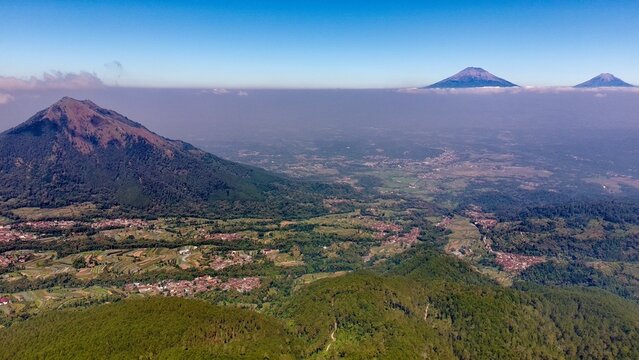 Aerial view of Mount Andong rising above lush green hills, terraced fields, and small villages under a clear blue sky in Ngablak, Central Java, Indonesia.