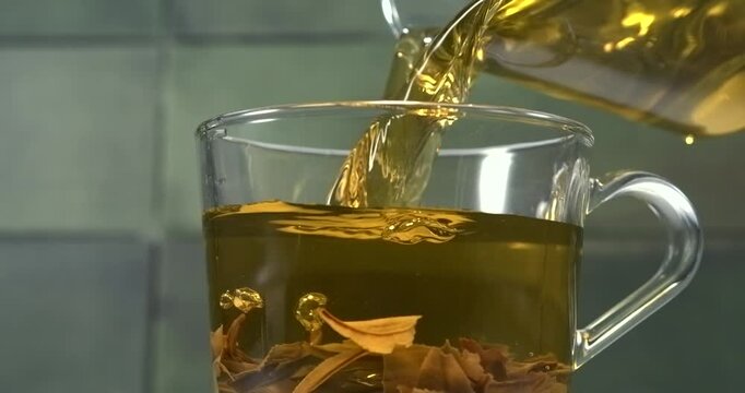 Hot green tea pouring into glass cup with tea leaves in slow motion