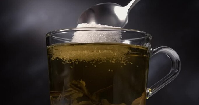 Sugar falling from spoon into hot green tea in glass cup in slow motion