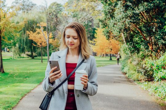 Smiling young woman using her phone while walking in autumn park for chatting, shopping, remote work, content creation for social media. Girl holding reusable cup with hot drink. and checking massages
