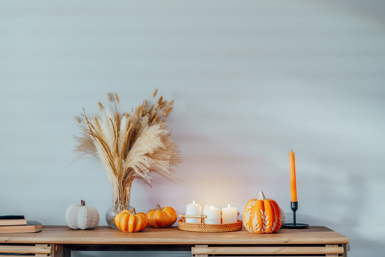 Cozy warm autumn home interior - decorative orange ceramic pumpkins, candles, dried flowers on the wooden console with white wall background. Scandinavian minimalist hygge home fall decor