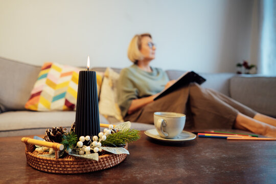 Focus on winter decor composition with burning candle and blurred woman planning goals for next new year while sitting on the sofa at home. Self-reflection, planning, action, checklist concept.