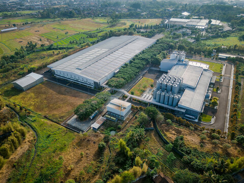 Aerial view of an industrial factory complex with a large warehouse and storage silos, surrounded by green rice paddies and rural landscape in Semarang Regency, Central Java, Indonesia.