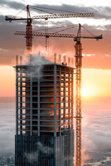 Obraz premium High angle view of a modern skyscraper under construction with tower cranes above a dense layer of white morning fog at golden sunrise, representing urban growth and business.