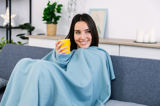 Young happy woman relaxing on sofa, wrapped in warm blanket, holding hot beverage, enjoying cozy winter day at home