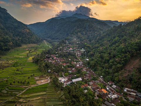 Aerial view of Kayuwangi Lor village nestled in a lush valley with vibrant green rice terraces and dense forest under a golden sunset sky in Banyubiru, Central Java, Indonesia.