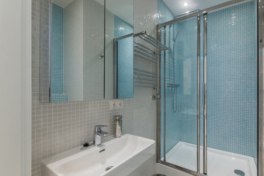 A small bathroom features blue tiles on the walls and a glass shower. A sink is present under a mirror, with modern fixtures. The space appears clean and organized