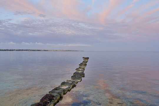 View of Saline dello Stagnone with a row of stones leading into the calm water under a pink sunset sky in Marsala, Sicilia, Italy.