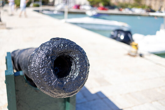 Close view of an old black harbour cannon in Zante Greece pointing towards the camera with boats and waterfront softly blurred behind creating a distinctive maritime detail