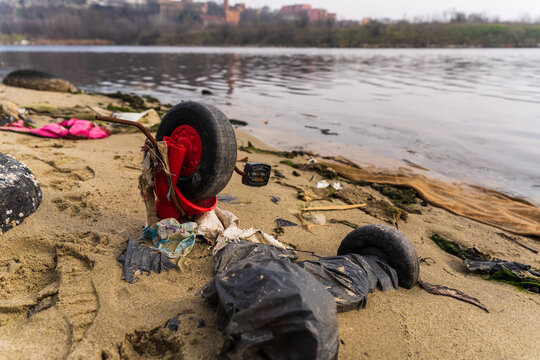 View of a discarded, broken toy bicycle on the sandy shore of Lake Varna, surrounded by plastic debris, with calm water and distant shoreline in the background in Varna, Bulgaria.