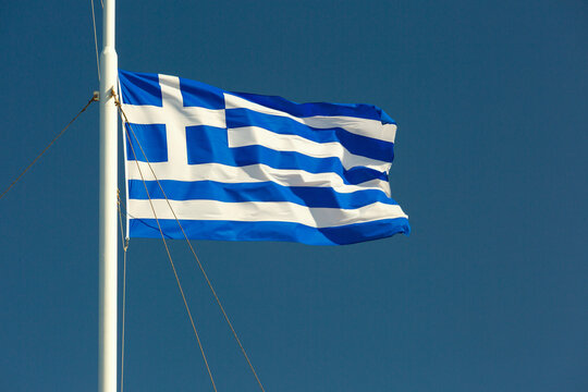 National flag of Greece waving on a flagpole against a clear deep blue sky.