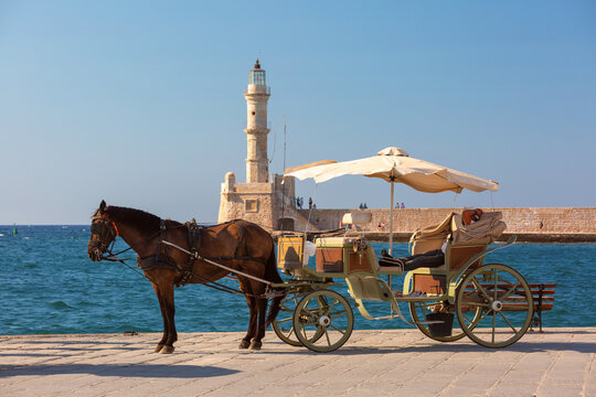 White horse with a traditional carriage on the waterfront overlooking the historic lighthouse of Chania Old Venetian Harbor in Crete, Greece.
