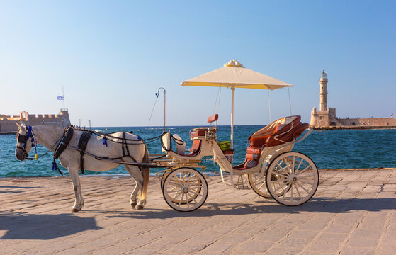 White horse with a traditional carriage on the waterfront overlooking the historic lighthouse of Chania Old Venetian Harbor in Crete, Greece.