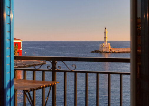 View through open balcony doors toward Chania Lighthouse, turquoise sea and boats in the Old Venetian Harbor of Chania, Crete, Greece.