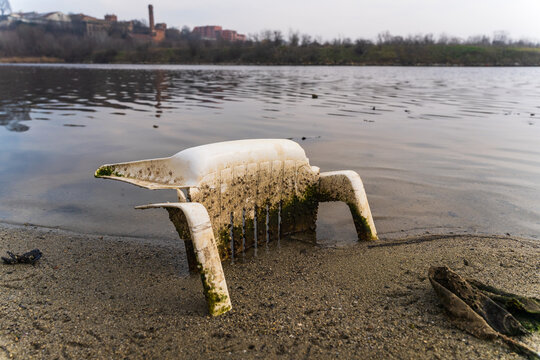 View of a discarded white plastic chair partially submerged in the water at the edge of Lake Varna, with algae growth on the frame and a sandy shoreline in Varna, Bulgaria.