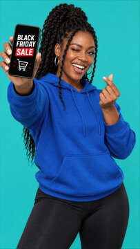 Happy Black woman in blue hoodie winking and making finger heart while showing Black Friday Sale on smartphone screen against turquoise background