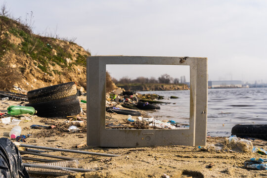 View of an old, empty television frame placed on a polluted beach with scattered tires and plastic debris along the shore of Lake Varna, Varna, Bulgaria.