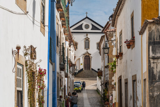 Obidos, Portugal - 13 May 2026: View of a narrow cobblestone street in the historic town of Obidos, featuring traditional white-washed buildings, flower pots, and a church at the end of the path.