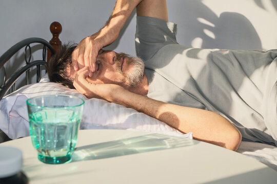 Man lying in bed suffering from a headache, covering his eyes from bright sunlight